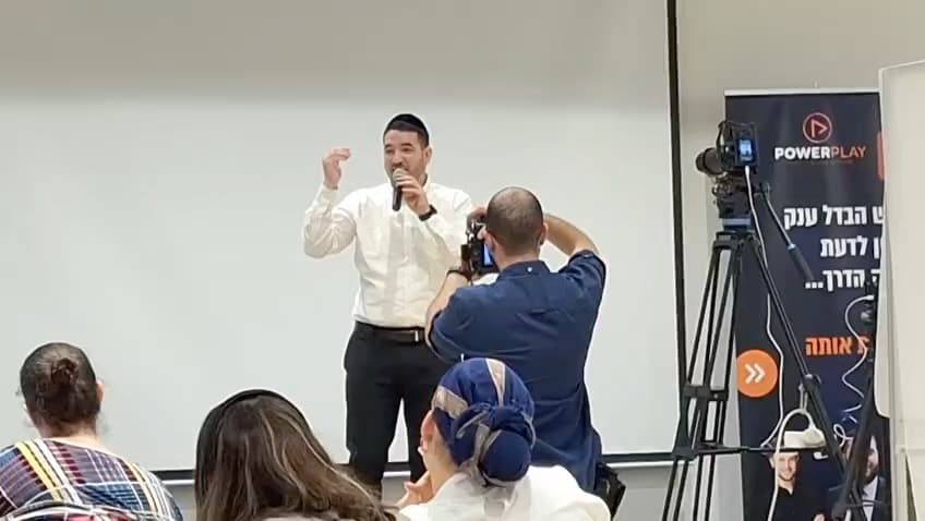 יוסף בן שושן מדבר אל הקהל ומסביר בתנועת ידיים, צלם מתעד מקדימה